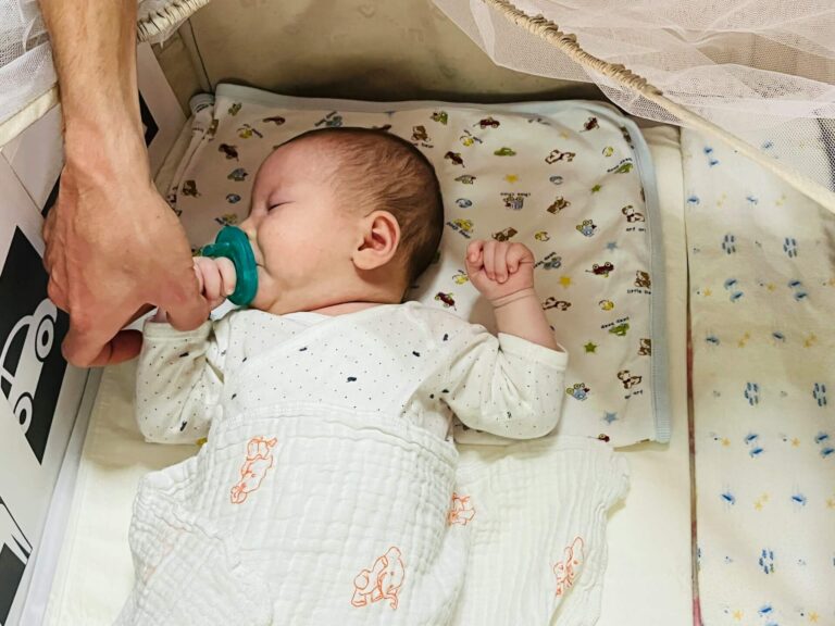 Baby sleeping in bassinet with mum's hand nearby